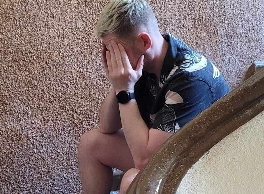 Man sitting on a staircase with peach wall behind him, holding his face in his hands and looking upset and distressed.